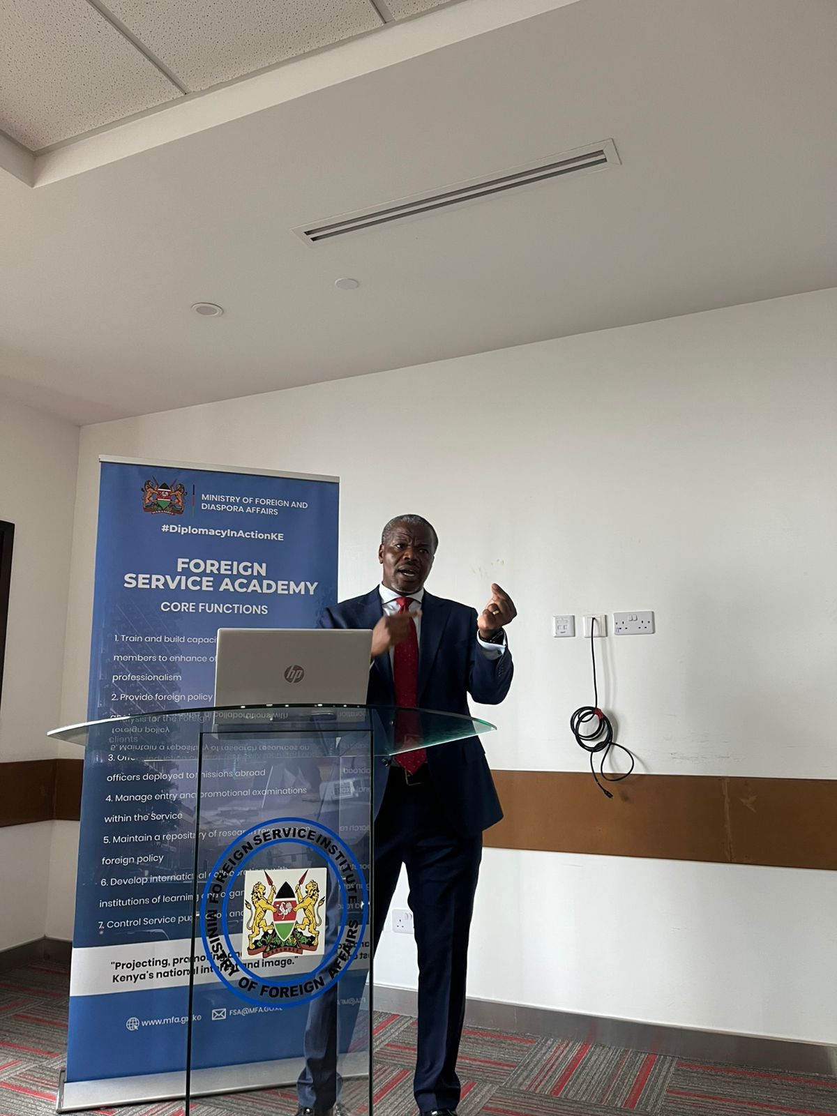 Dr. James Nyawo lecturing at a Foreign Service Academy podium beside a Diplomatic Training and Research banner